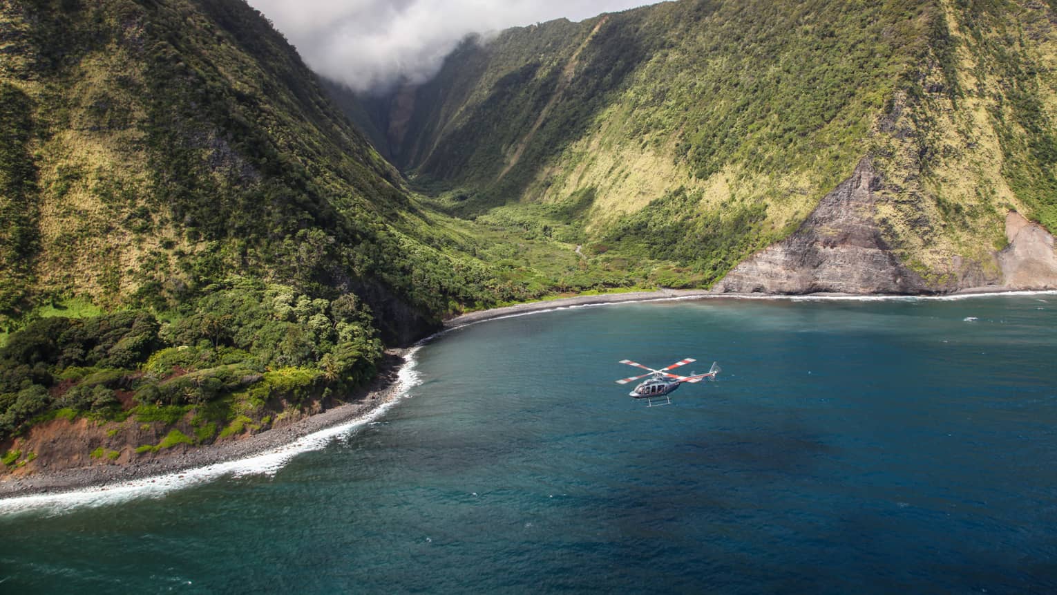 Helicopter flies over ocean by sweeping green volcanic mountains