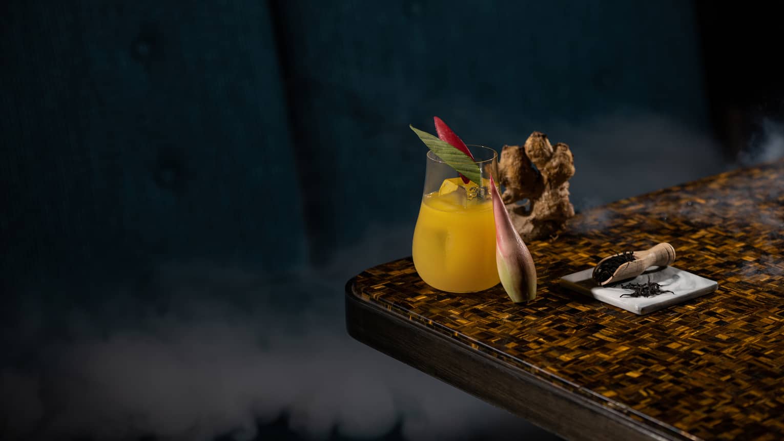 Kunyit Royale signature cocktail in stemless glass on edge of countertop with turmeric root and leaf garnish
