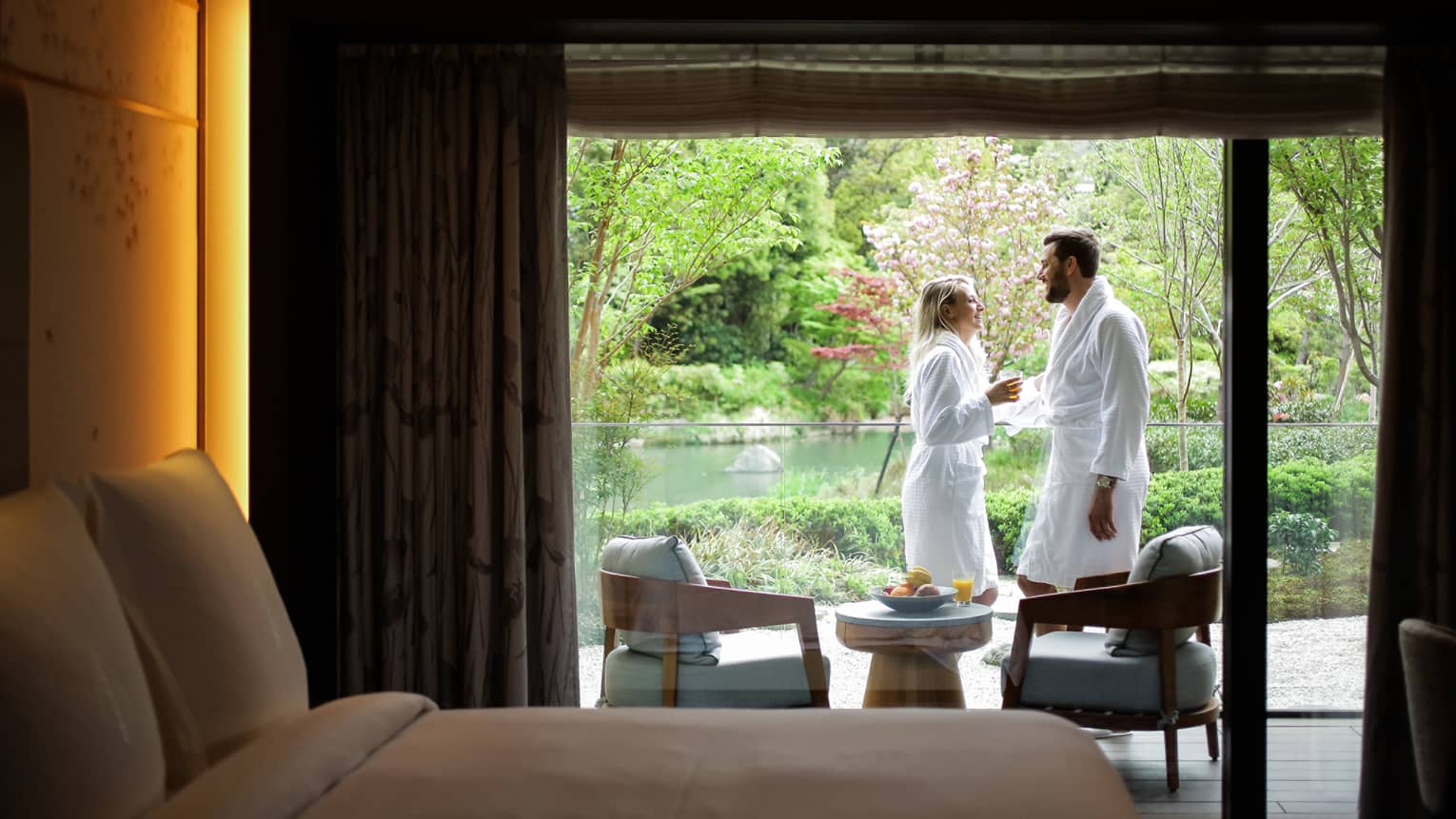 Smiling couple wearing white bathrobes on sunny balcony past hotel room bed