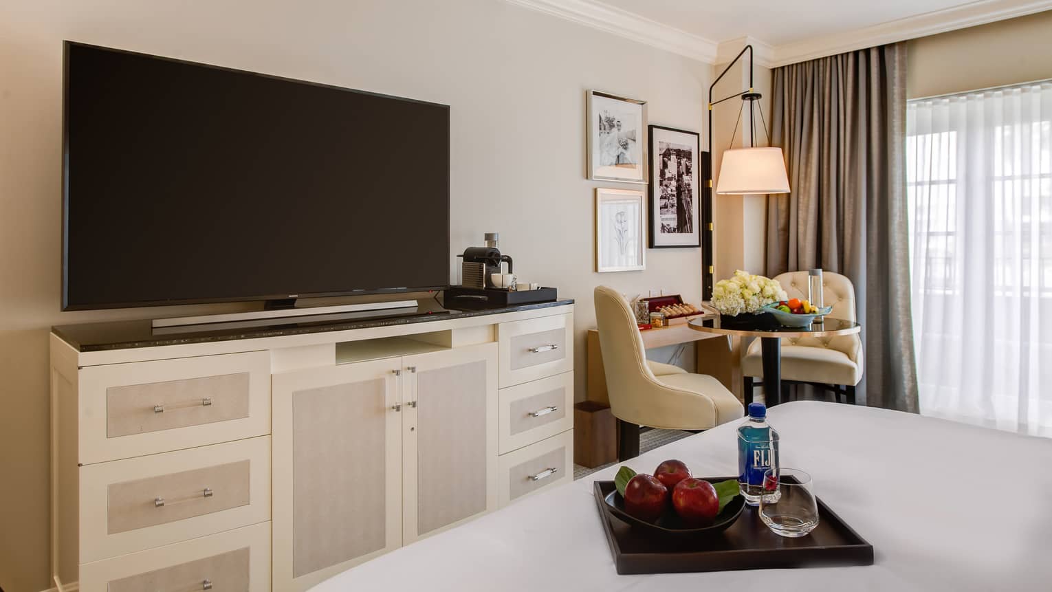 Large TV on dresser by foot of hotel bed with wood tray, red apples and Fiji water bottle