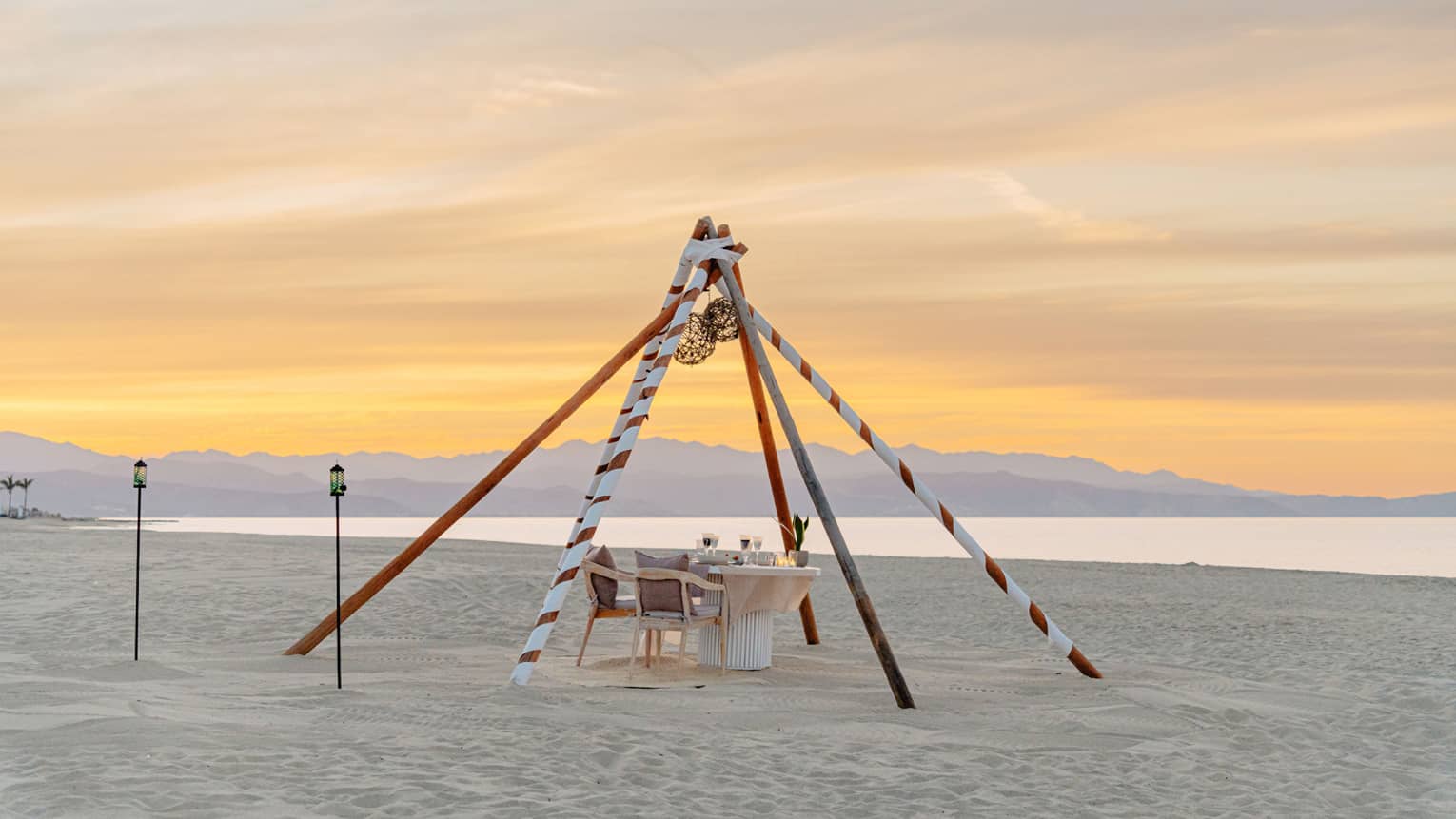 A dining table with seating and lighting on the beach at sunset
