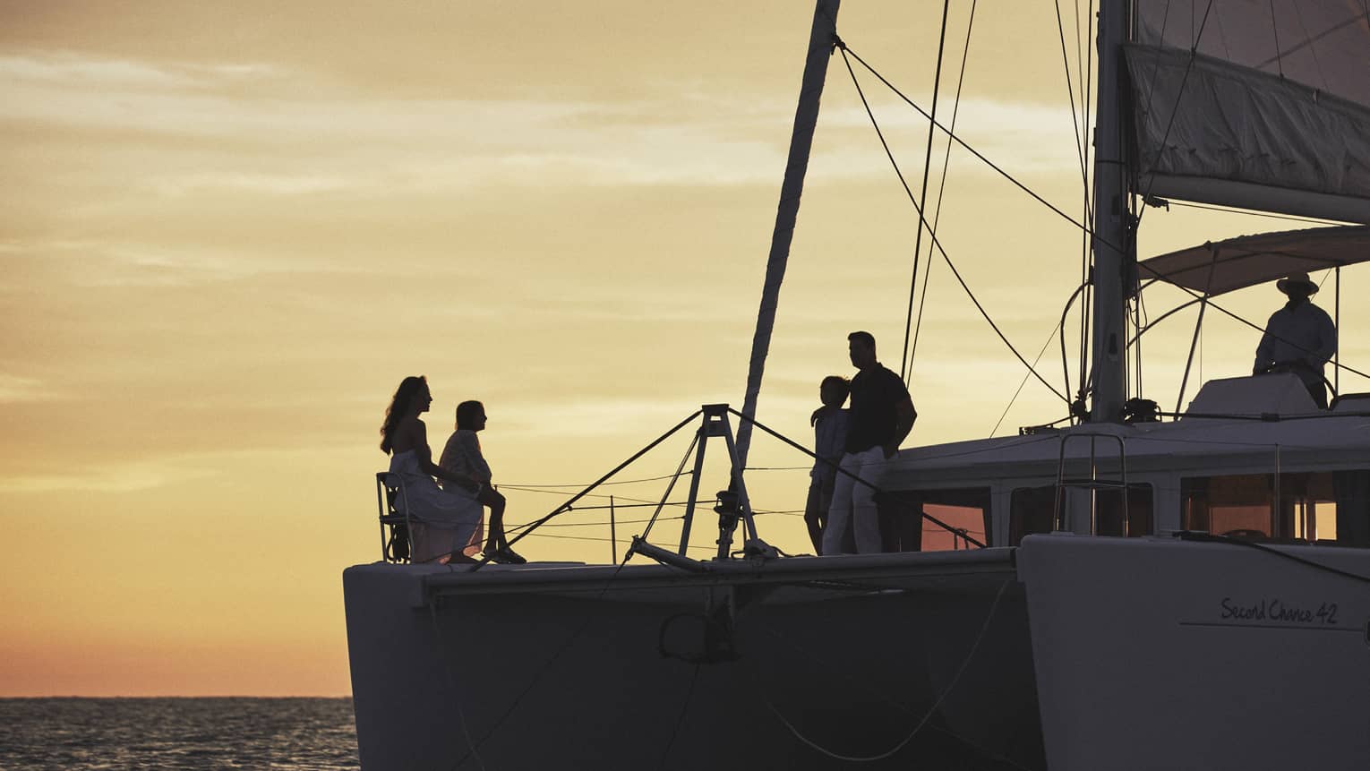 A family gathers at the bow of a large sailboat, silhouetted under a golden evening sky, captain steering from the cockpit.