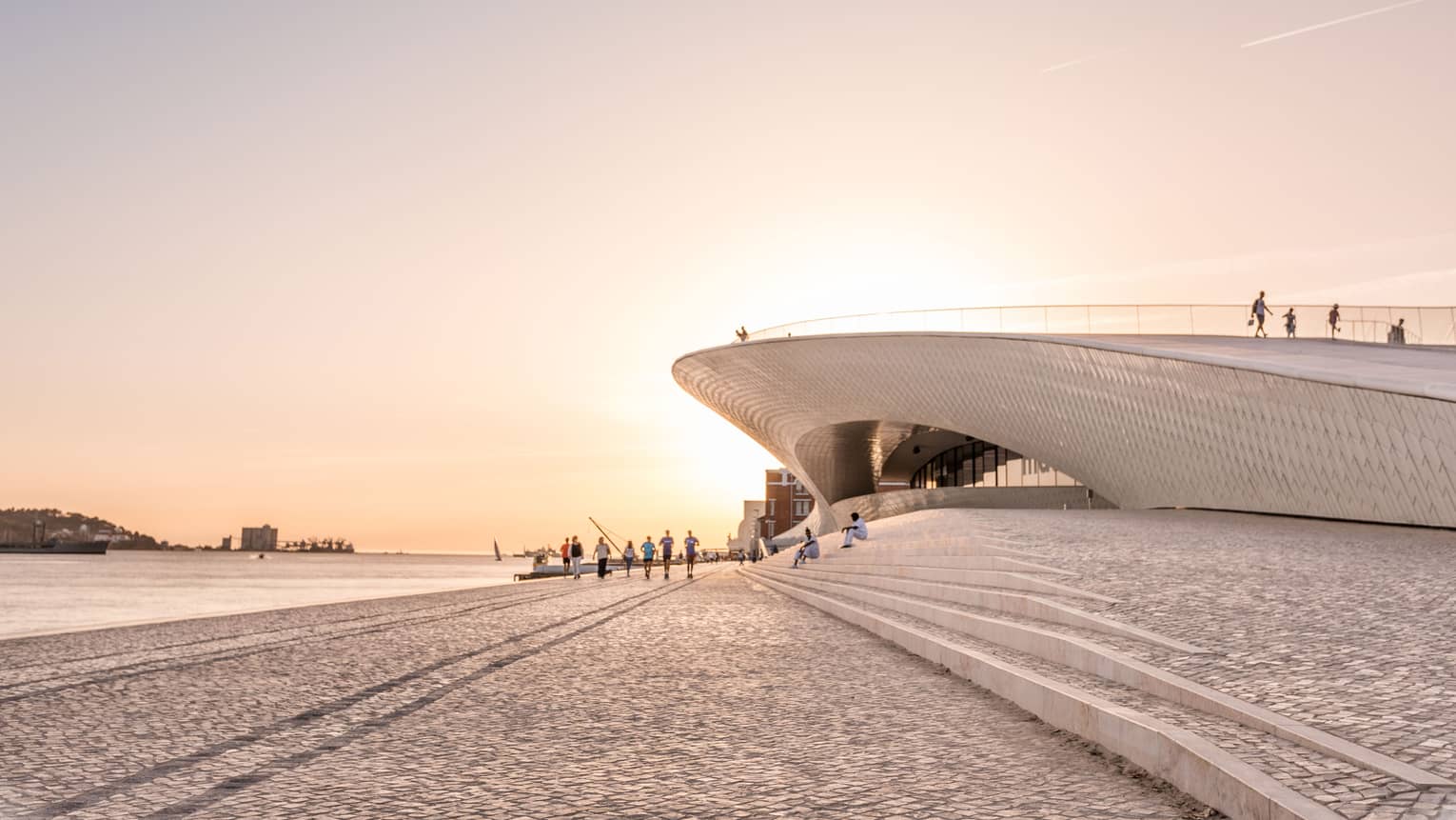 Tourists walk by a disc-shaped modern building next to a harbour. The setting sun reflects off the cobbled walkway. 