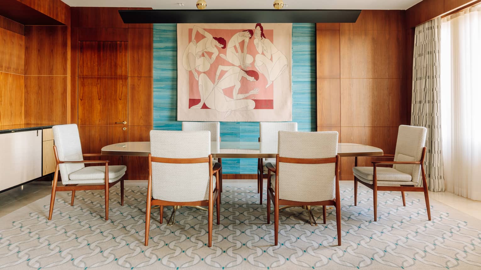Dining room with a long table, six chairs, colorful abstract wall art and wood-paneled walls with natural light from large windows.
