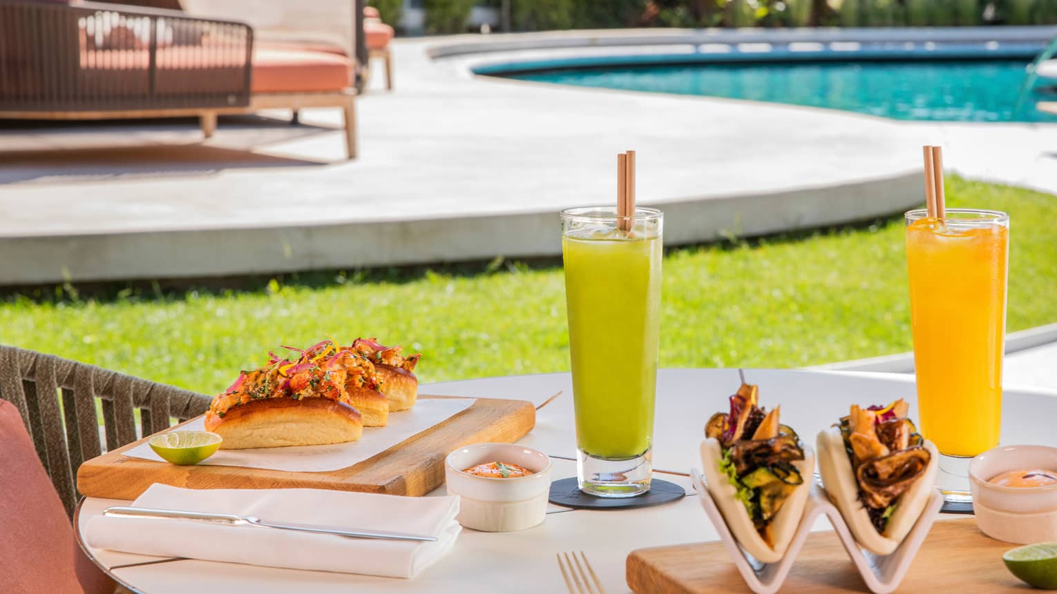 Poolside table plated with tall cocktails and lobster rolls on boards