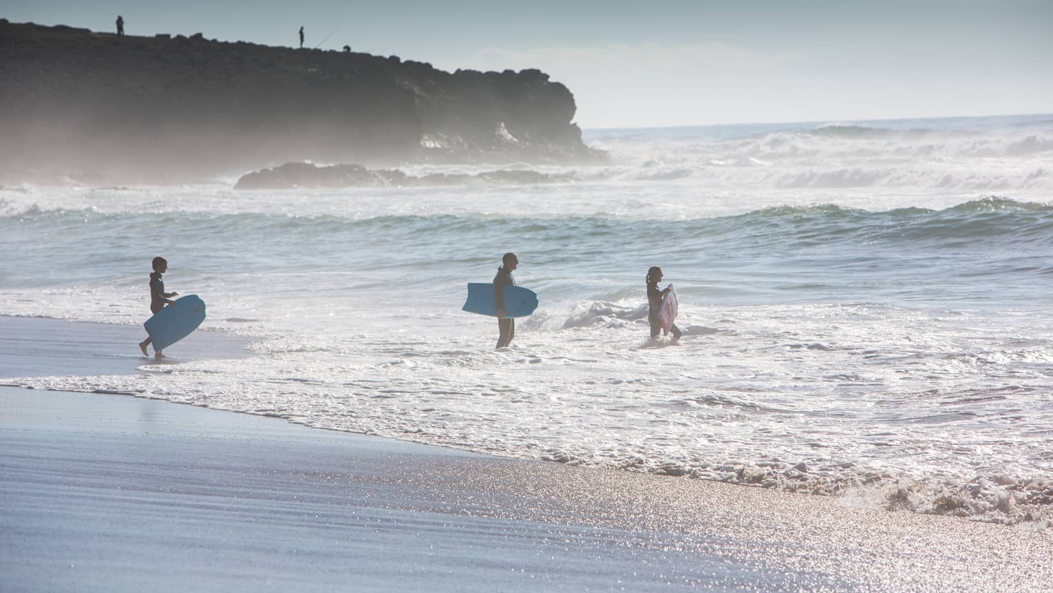 Silhouettes of man, woman and child all wearing wetsuits, holding blue surfboards, walking into ocean