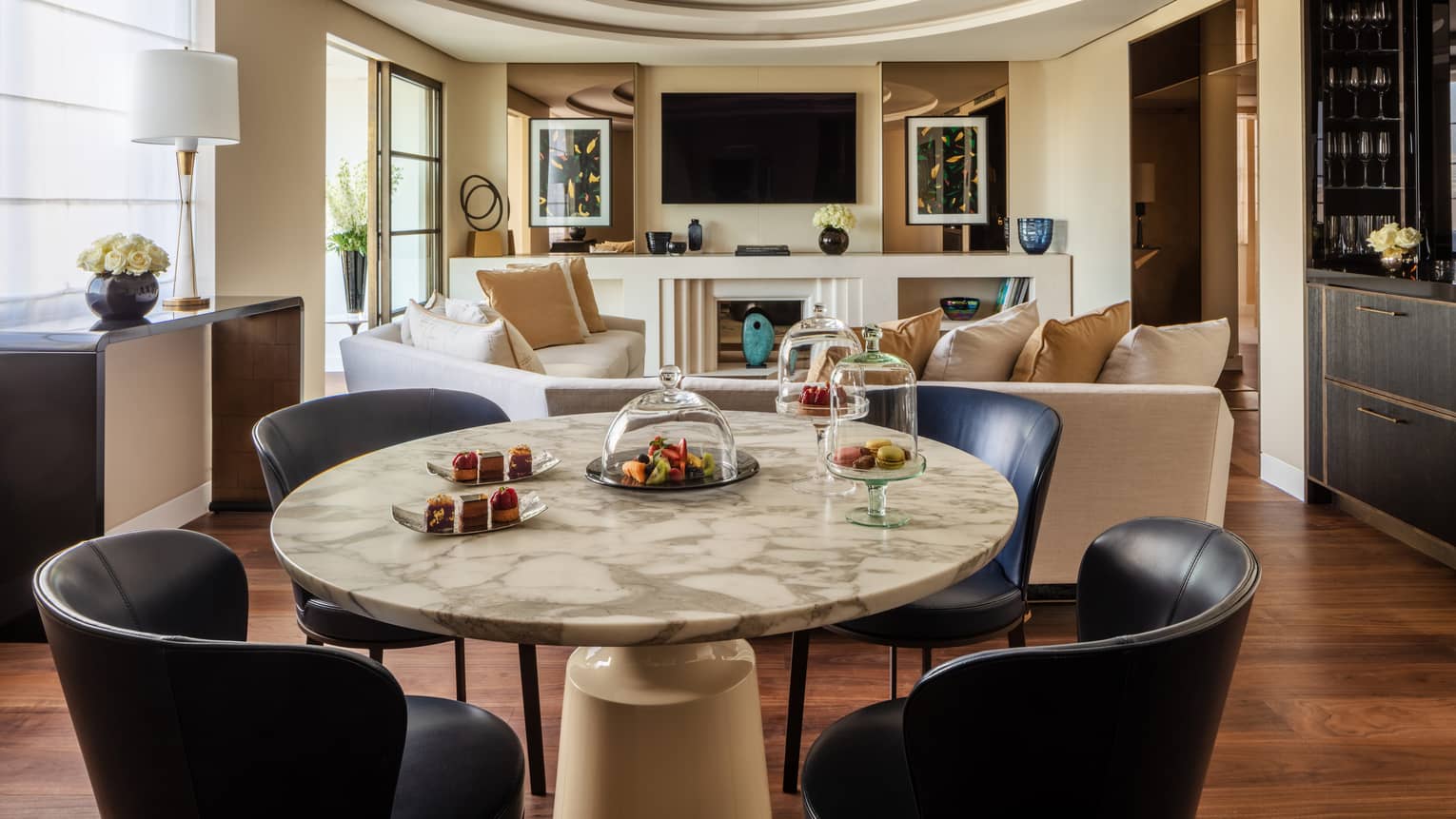 A suite with a round stone table with black chairs, a beige sofa, a television and large windows.