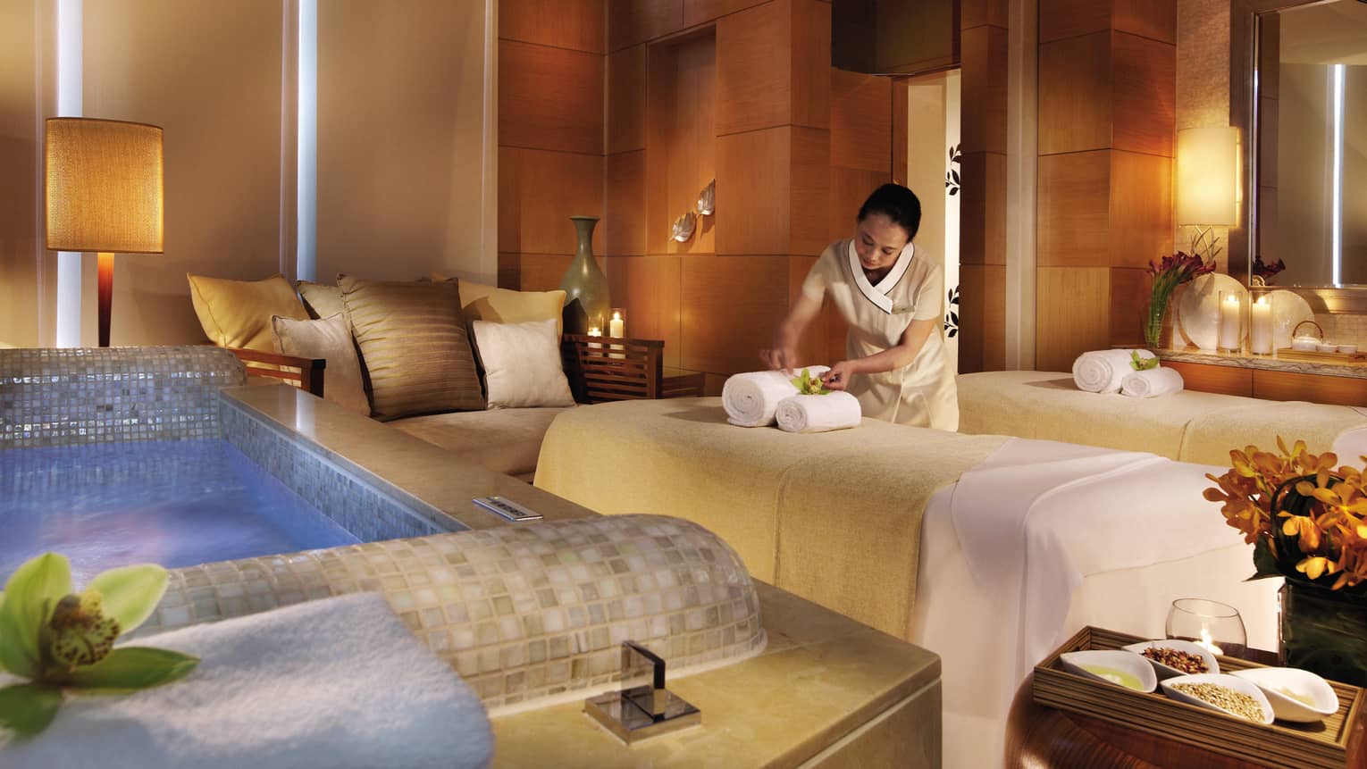 Spa staff sets rolled towels on massage bed near small sofa, tile bathtub in Couples Suite