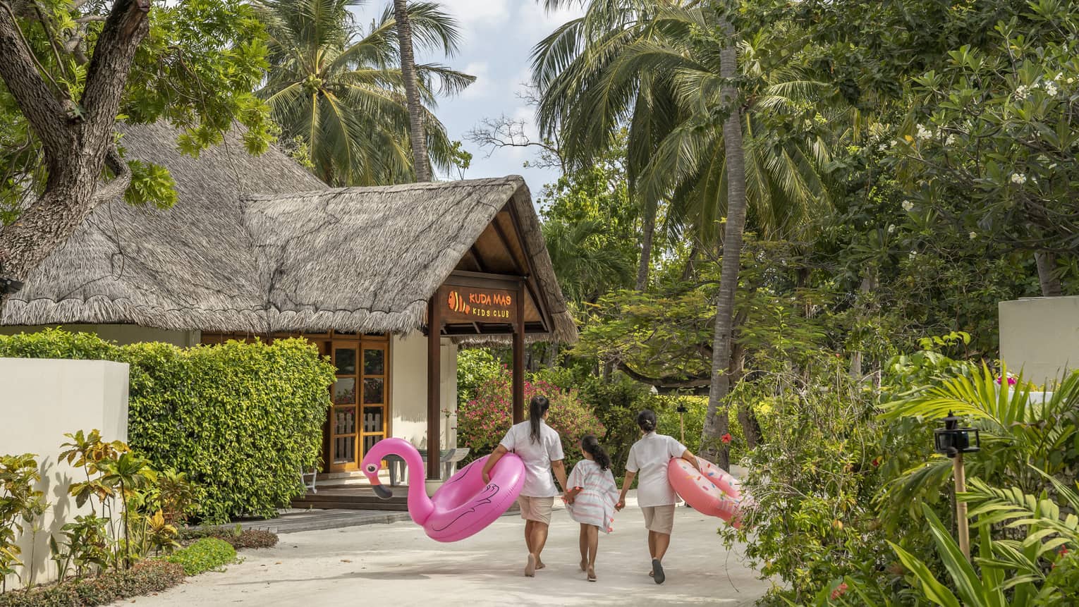 ,Two staff members holding flamingo pool flotation devices walk along tree-line pathway to thatched-roof pavilion