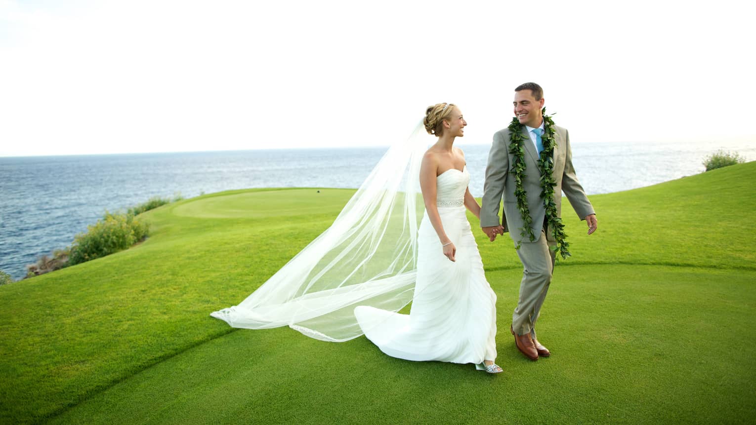 Bride and groom hold hands, walk along green lawn overlooking ocean