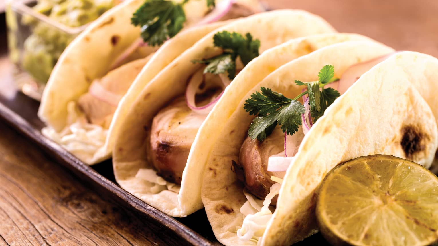 Row of three fish tacos, guacamole, lime on tray