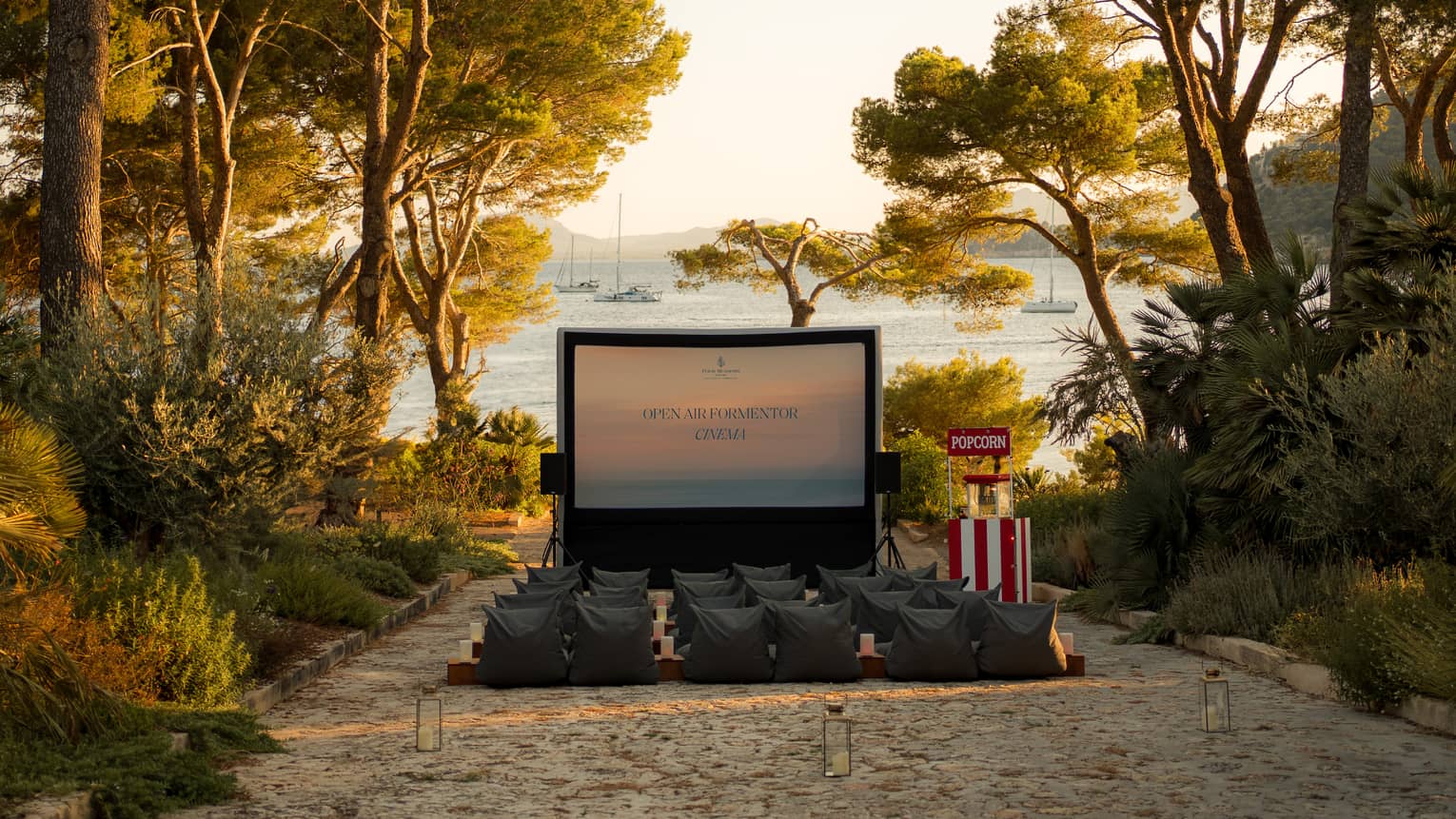 A large movie screen and rows of chairs in an outdoor setting  in the daytime. The movie area is surrounded by trees, and the ocean is in the background.