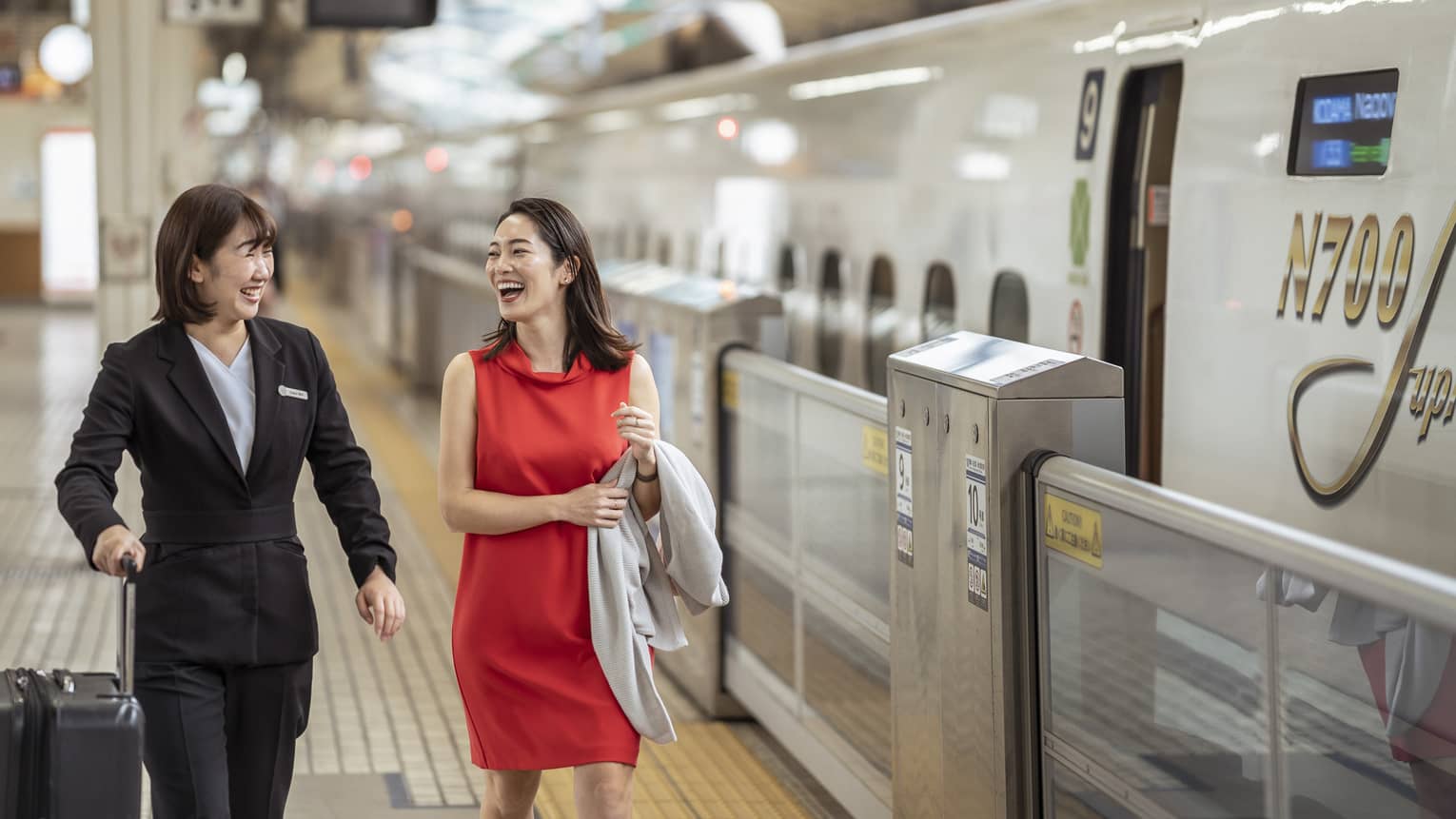 A Four Seasons employee holds a traveller's suitcase; laughing, they walk along the platform beside an N700 high-speed train.