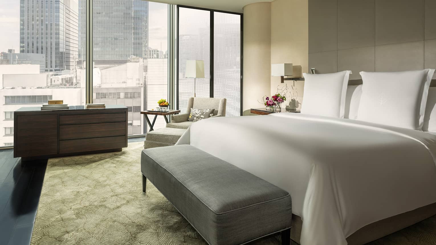 Modern hotel room with a king bed, grey tufted bench, cozy seating area, dresser and large windows offering city views