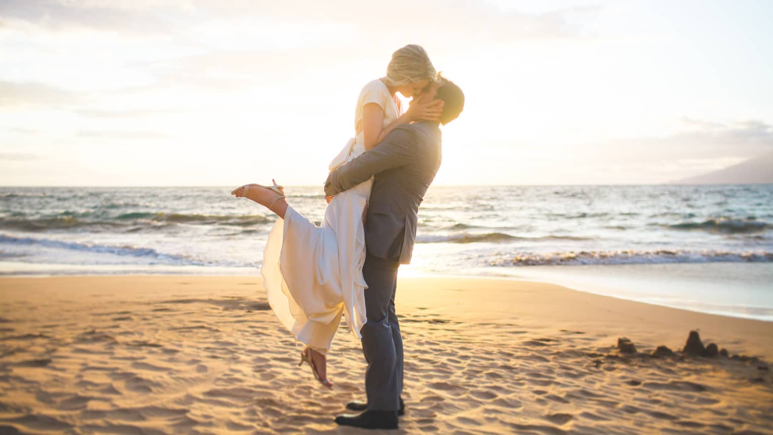 Groom lifts bride off ground, kiss on sand beach at sunset