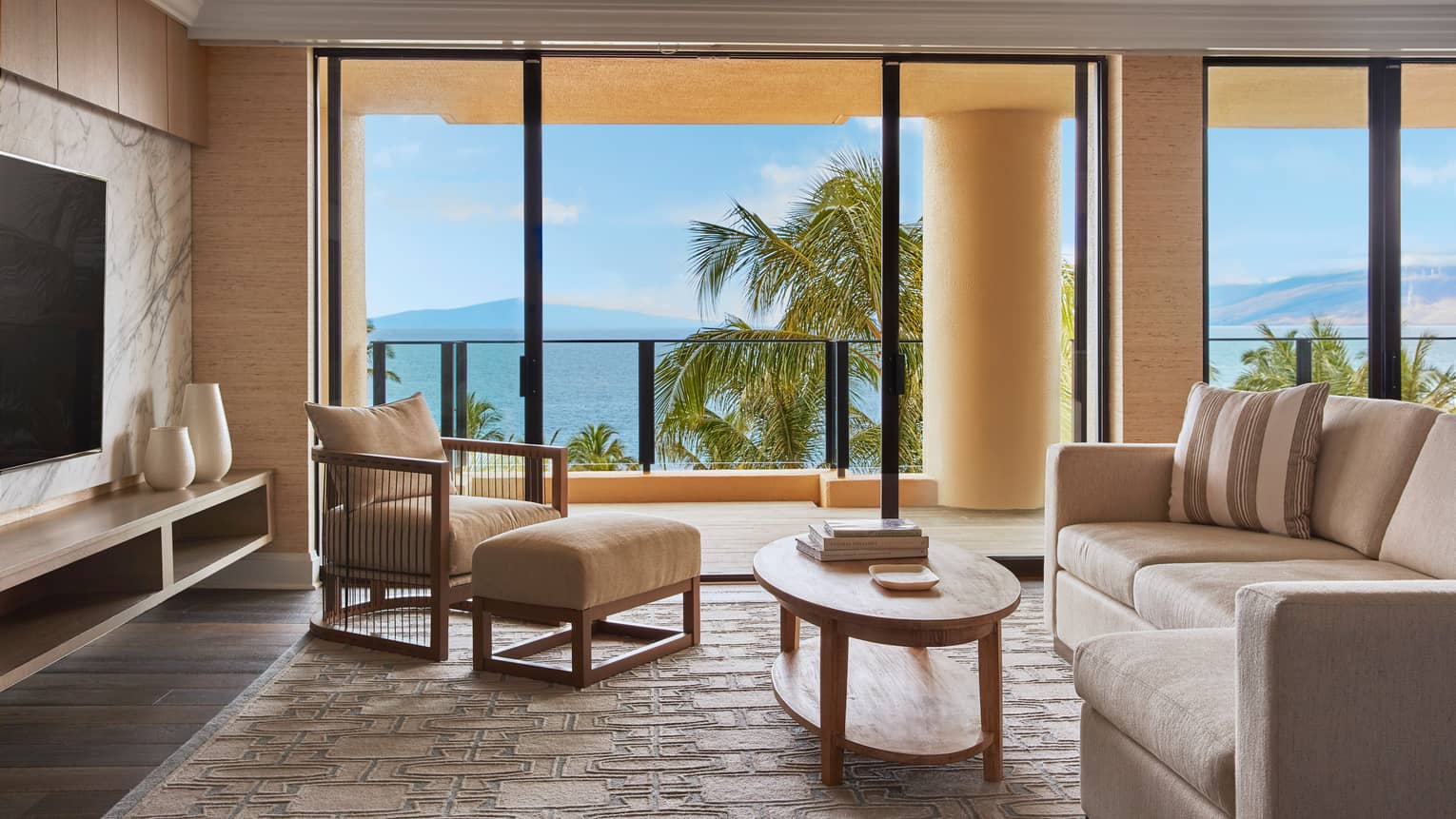 Living room with beige sofa, armchair and wooden coffee table, featuring floor-to-ceiling windows with ocean and palm tree views at a luxury resort suite