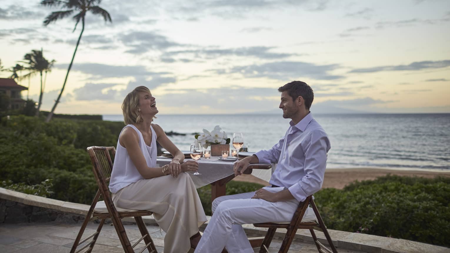 an in dress shirt and khaki pants, woman in white dress drink wine at outdoor cocktail table at sunset