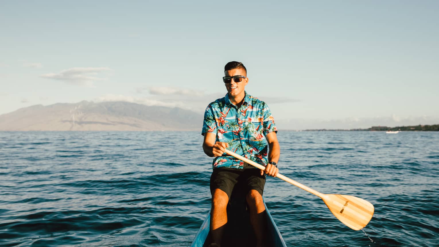 Person wearing a blue button-down patterned shirt, black shorts and sunglasses paddles through the water in an outrigger canoe