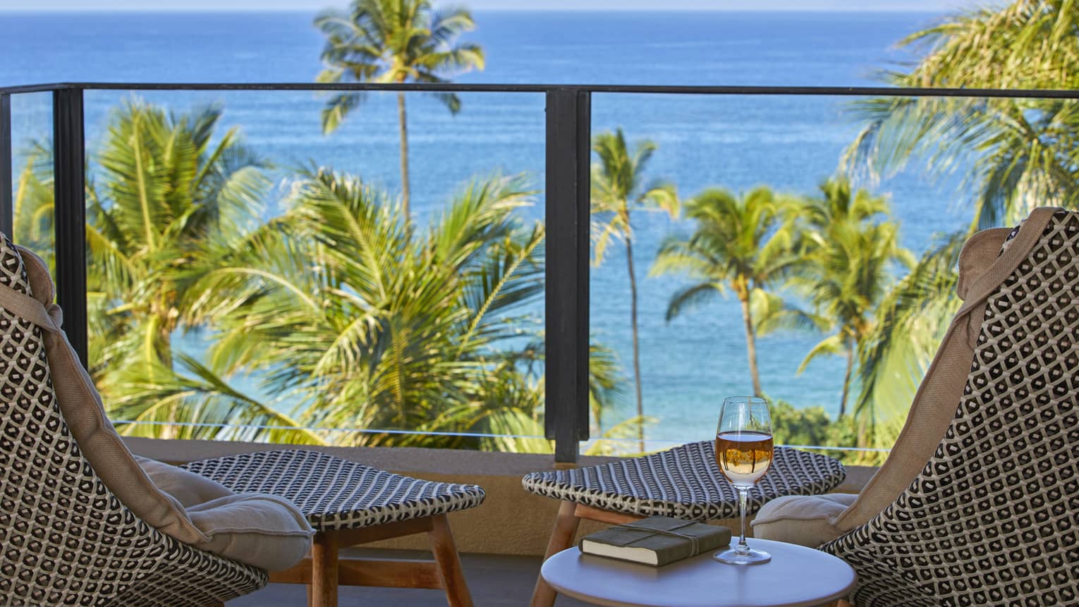Balcony with two patterned chairs, ocean view palm trees and a table with a book and glass of wine