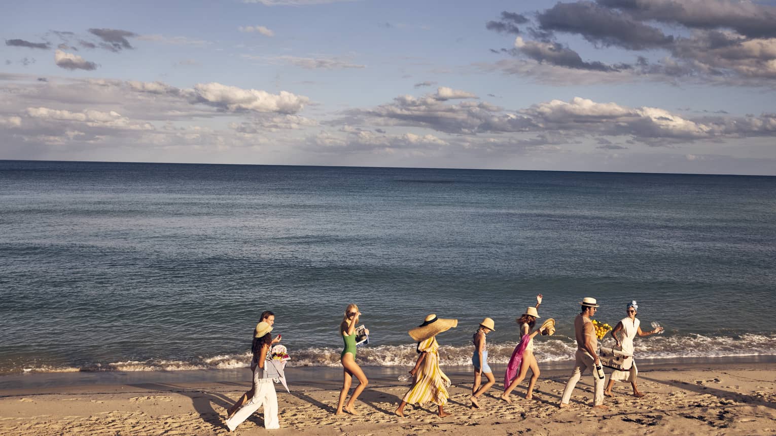 A group of people walking along a sandy beach by the ocean, wearing summer clothing and hats, with a backdrop of a calm sea and partly cloudy sky.