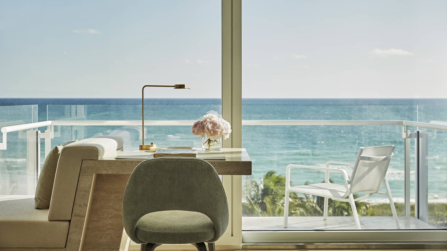 Luxury oceanfront hotel room desk with a chair, a lamp and pink flowers, overlooking a balcony with ocean views under a clear sky