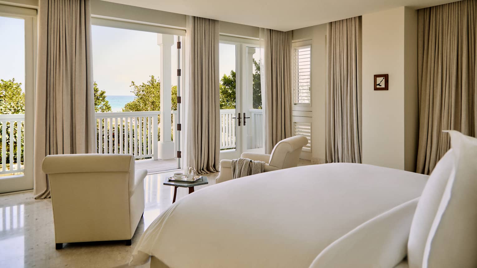 Luxury villa bedroom featuring a king bed and two beige arm chairs looking out to ocean view