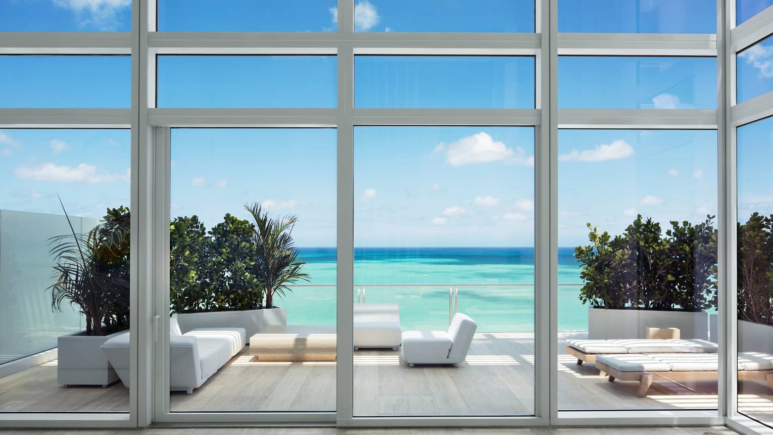 View from glass walls to large patio with modern white furniture, turquoise ocean