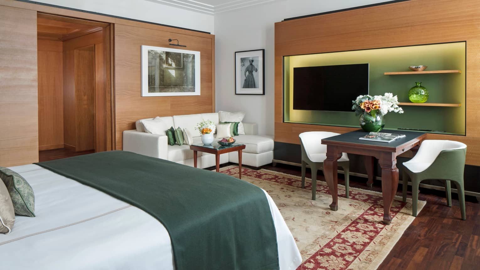 Junior Suite bed with emerald green blanket, wood panel wall with TV, small sofa, cafe table and chairs