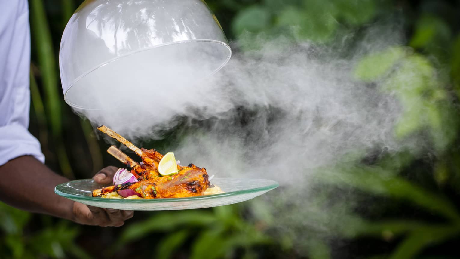A Four Seasons dining staff taking the lid off of a dinner tray – steam is pouring out over a cooked meat dish on skewers