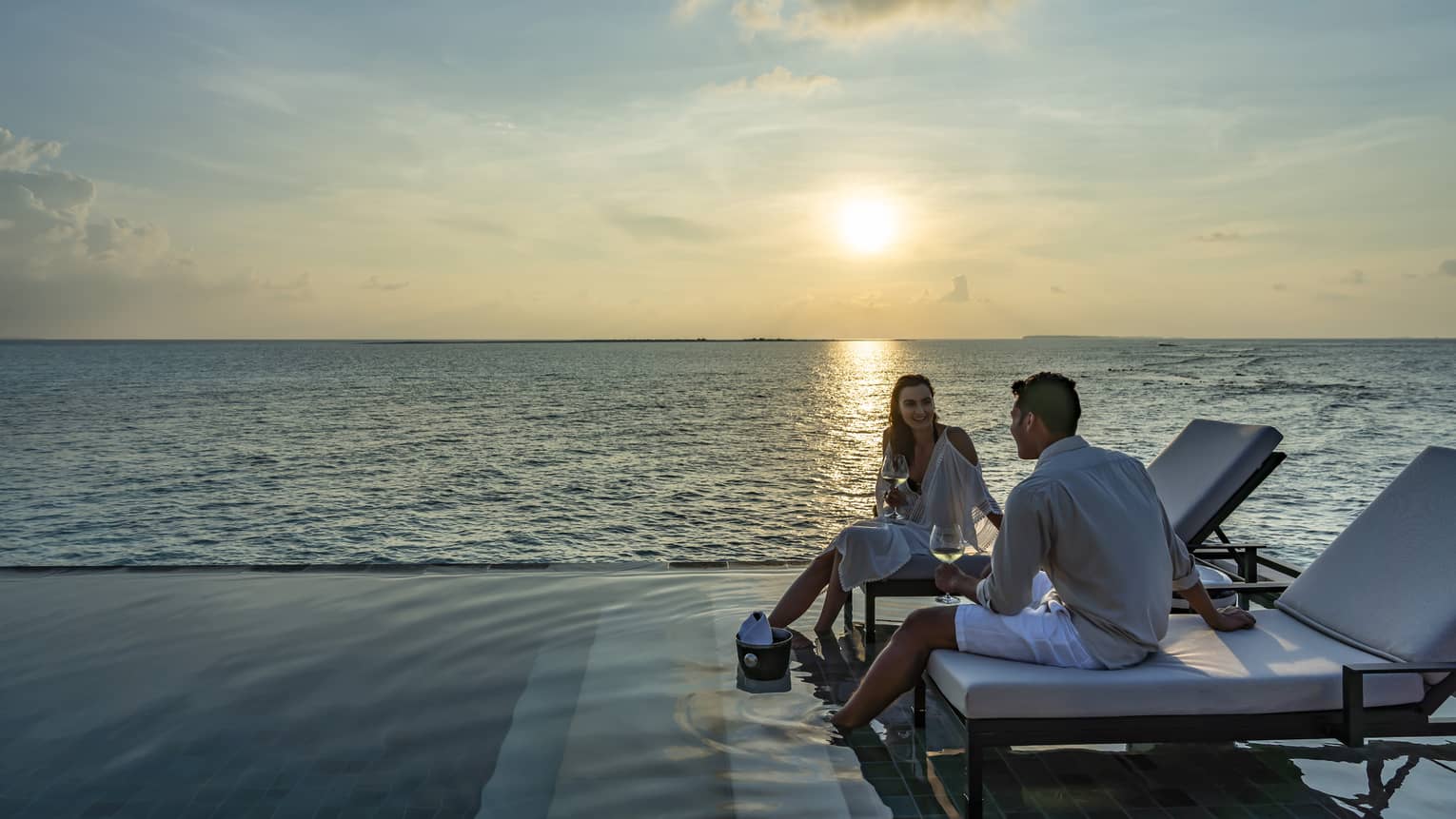 Couple hold wine glasses, dip feet in outdoor swimming pool by ocean at sunset