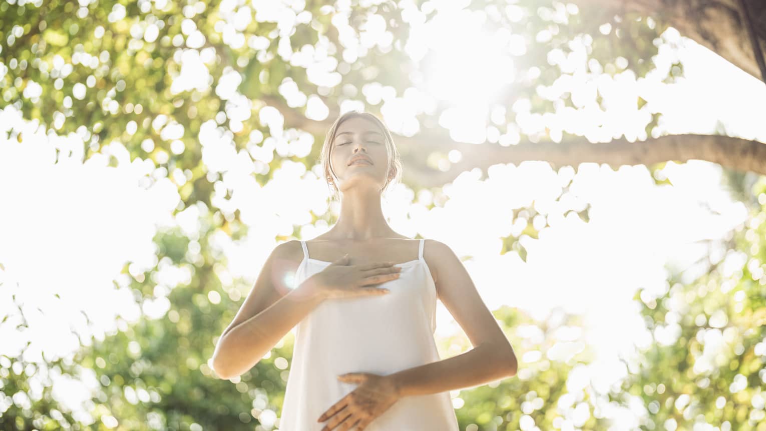 Person practicing mindful breathing in a wellness haven, standing under sunlit trees with hands on chest and abdomen