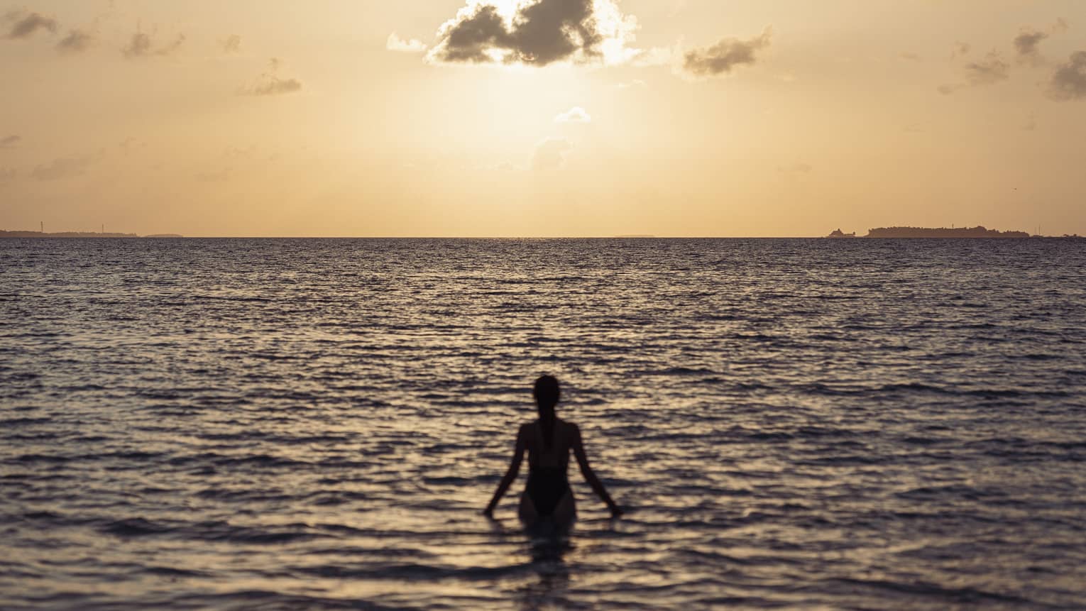 Woman silhouetted by the setting sun walks out into the ocean