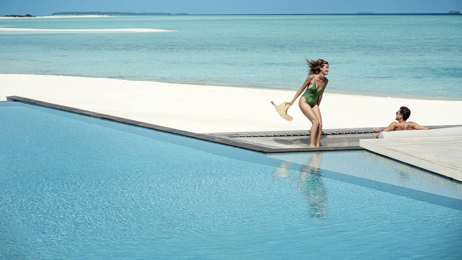 Woman and green one-piece swimsuit relaxes poolside with her male partner in the infinity pool, the vast blue waters of the lagoon behind them