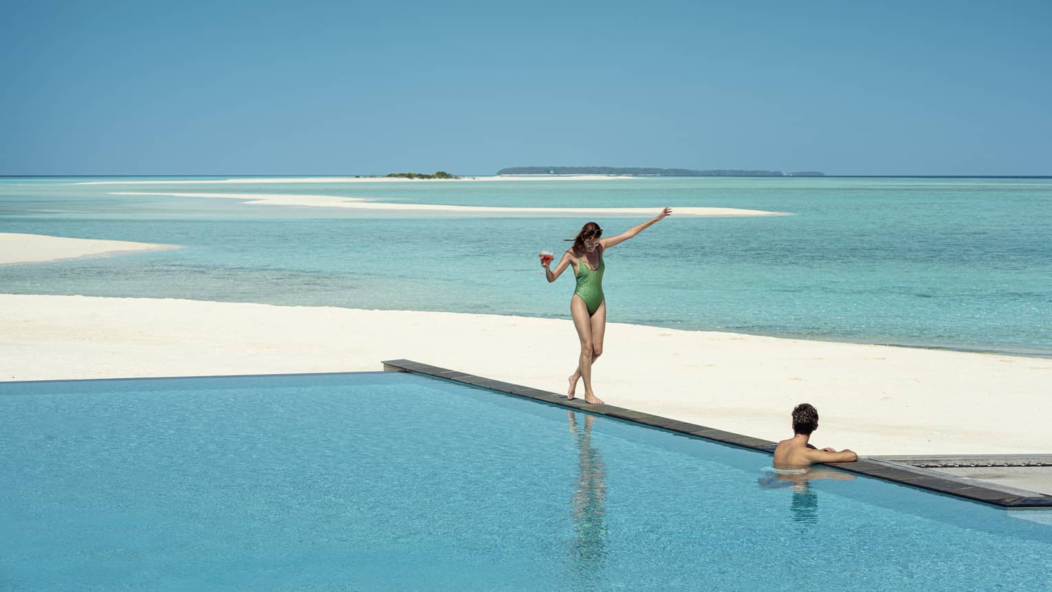 A guest wades in a pool on a beach overlooking the ocean as another guest balances on the edge with a drink in hand.