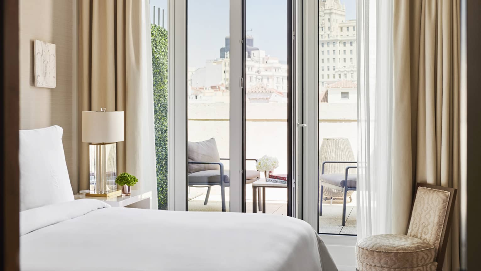 Luxury hotel bedroom with a king bed, small armchair, bedside table with lamp and a view of an outdoor terrace with seating through glass doors