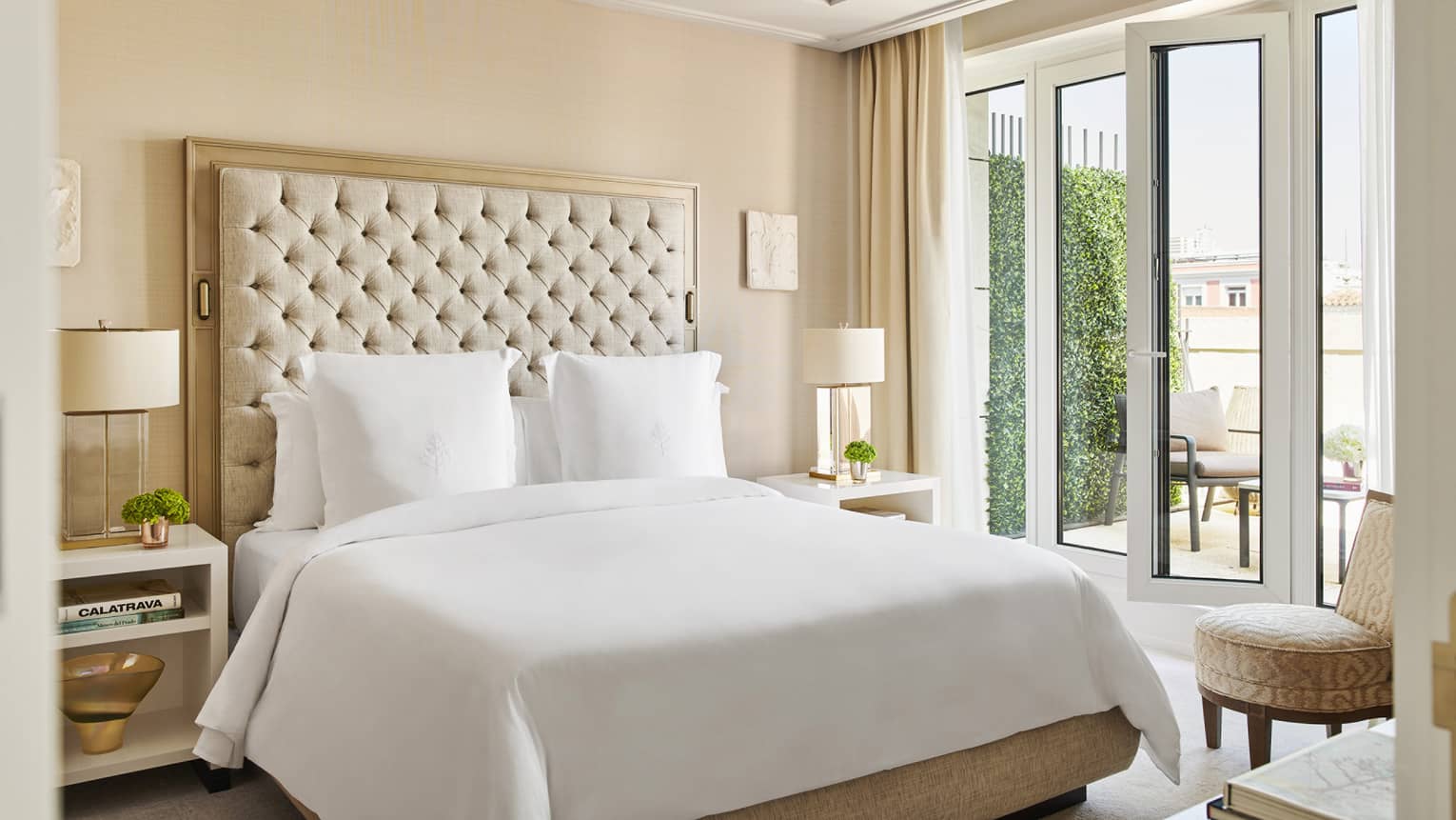 Luxury hotel bedroom with a king bed, tufted headboard, bedside tables with lamps and French doors opening to a private terrace with outdoor seating