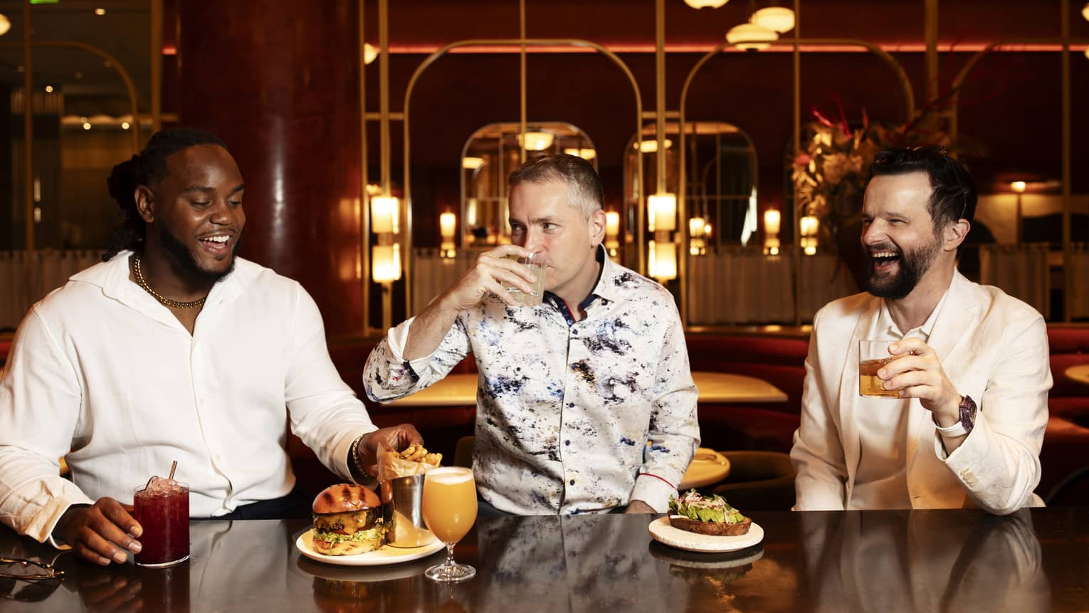 Three guests sitting at the bar with food and drinks, with lights in the background.