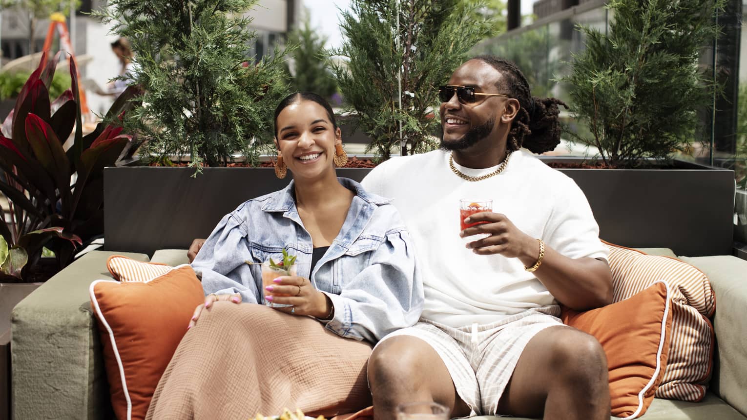 Two guests at an outdoor terrace sitting on a couch with orange pillows and holding drinks.