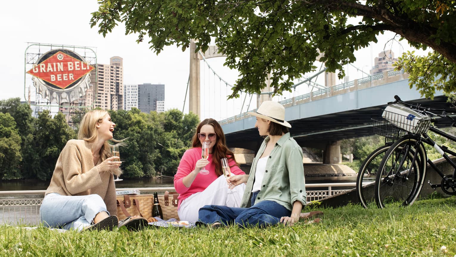 Three women have a picnic in a city park and toast with champagne