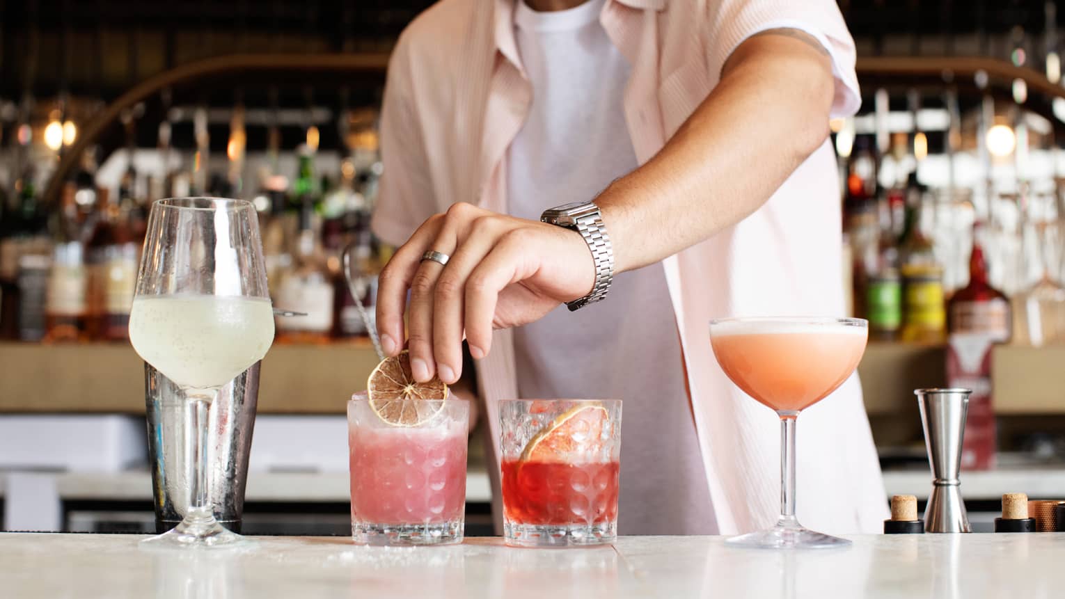 Bartender wearing light-pink short-sleeve shirt over a white t-shirt places garnishes on a row of row brightly coloured cocktails