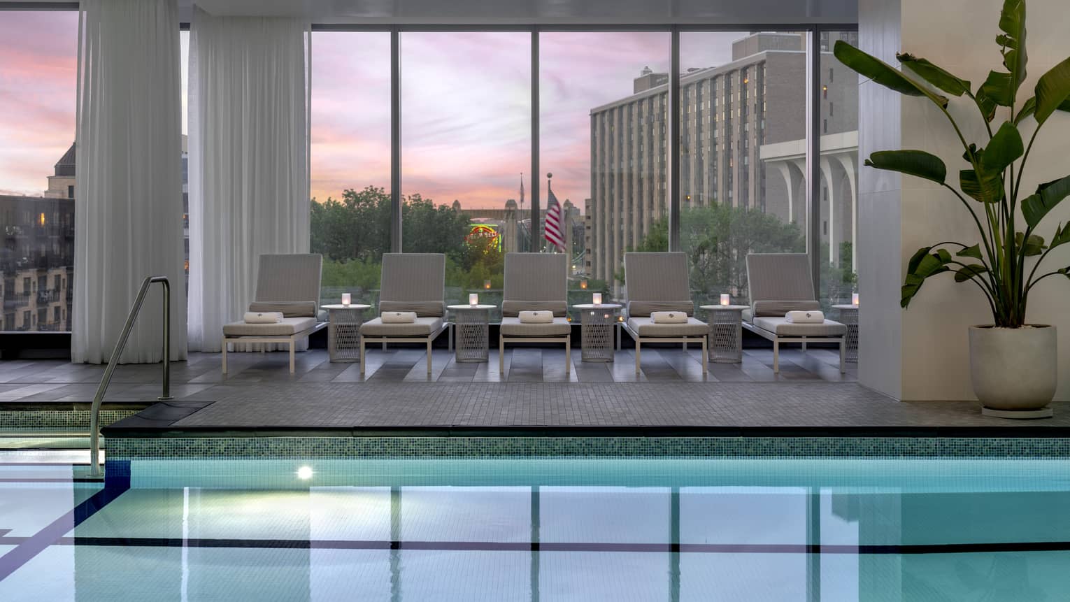 An indoor pool with dim lighting, plants, and lounge chairs with towels.