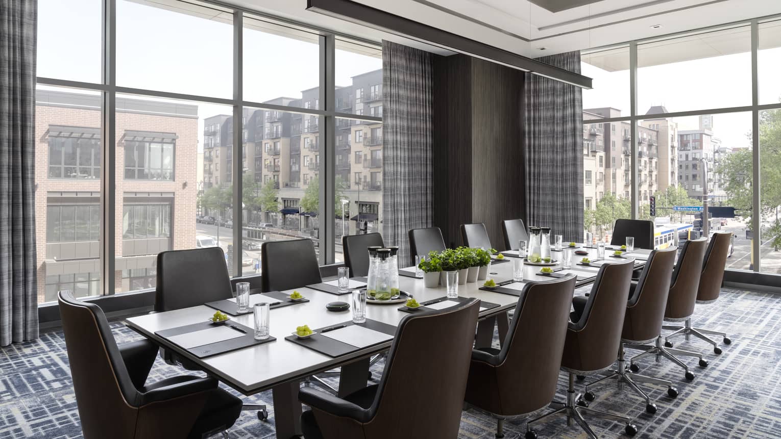 A meeting room with large windows, a long table, brown chairs and papers and supplies on table.