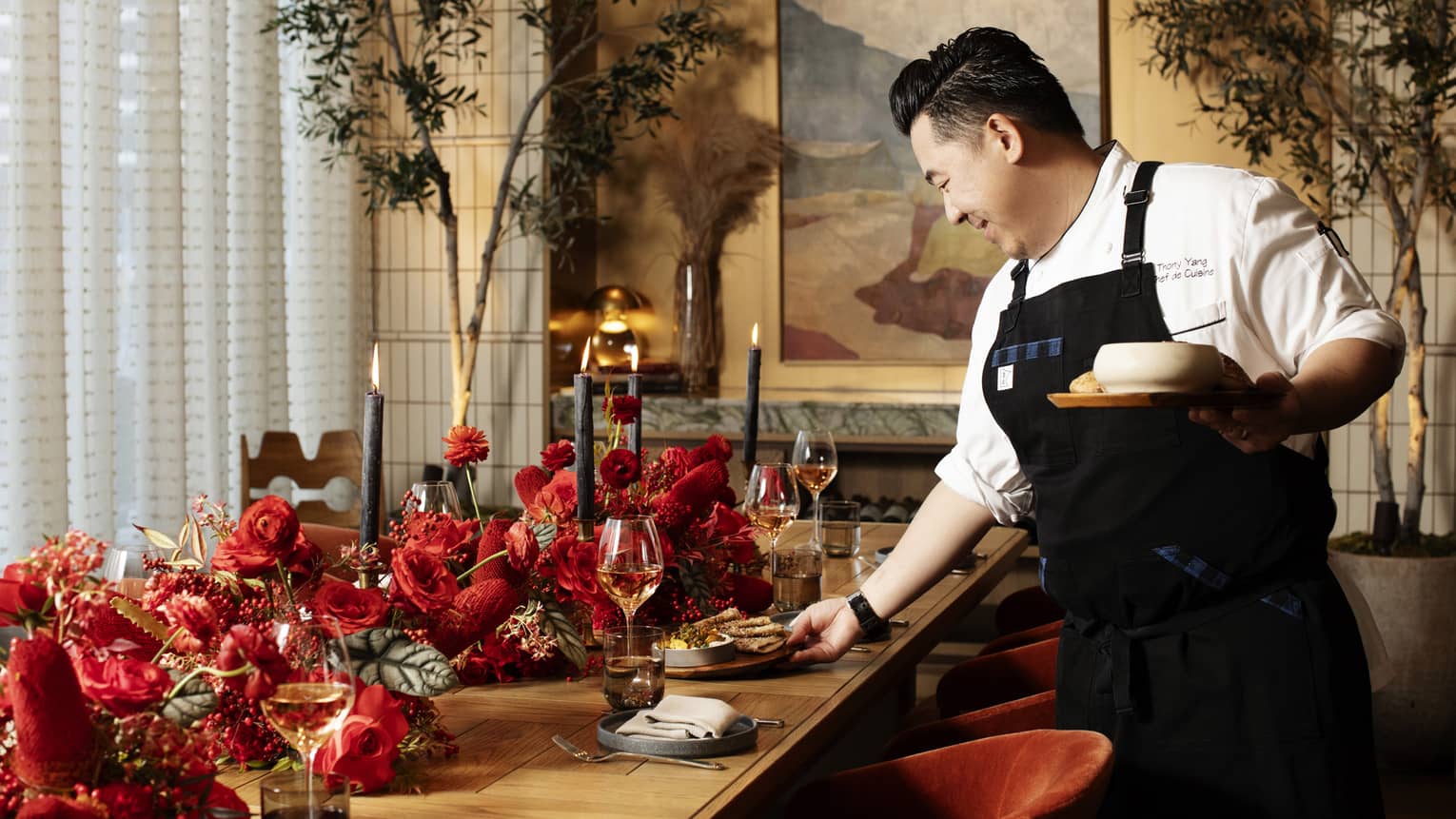 Chef putting dishes on a table with red flowers on it.