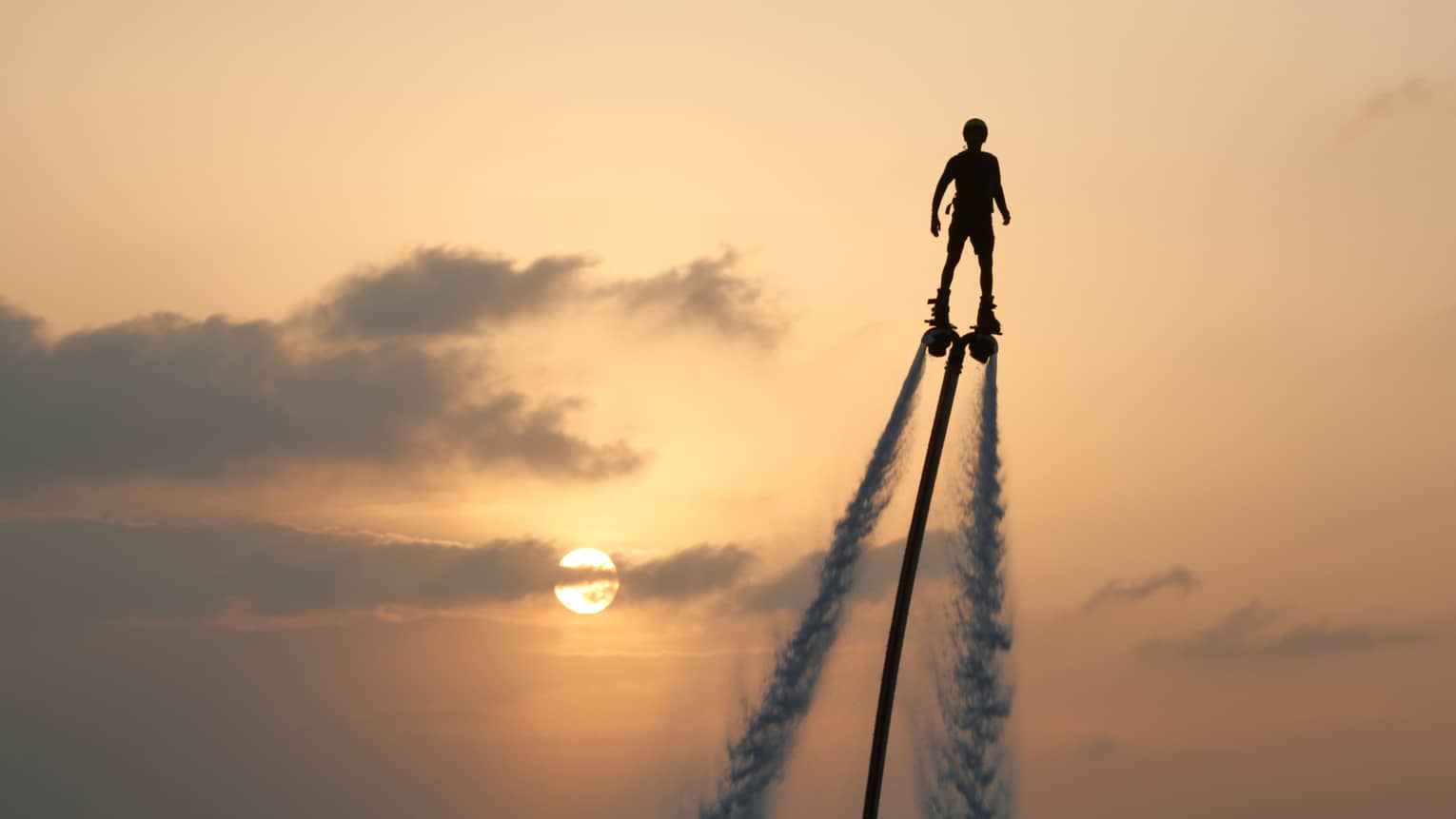 Silhouette of person soaring high above ocean on jet pack, water trails below, against sunset