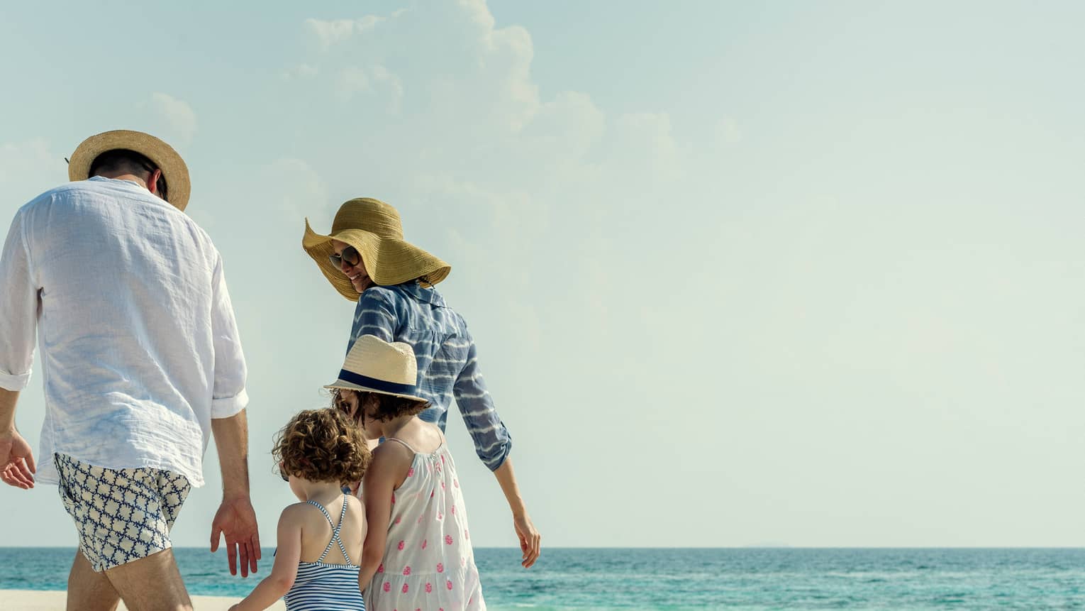 Family wearing swimsuits, sundresses and sun hats walk along white sand beach