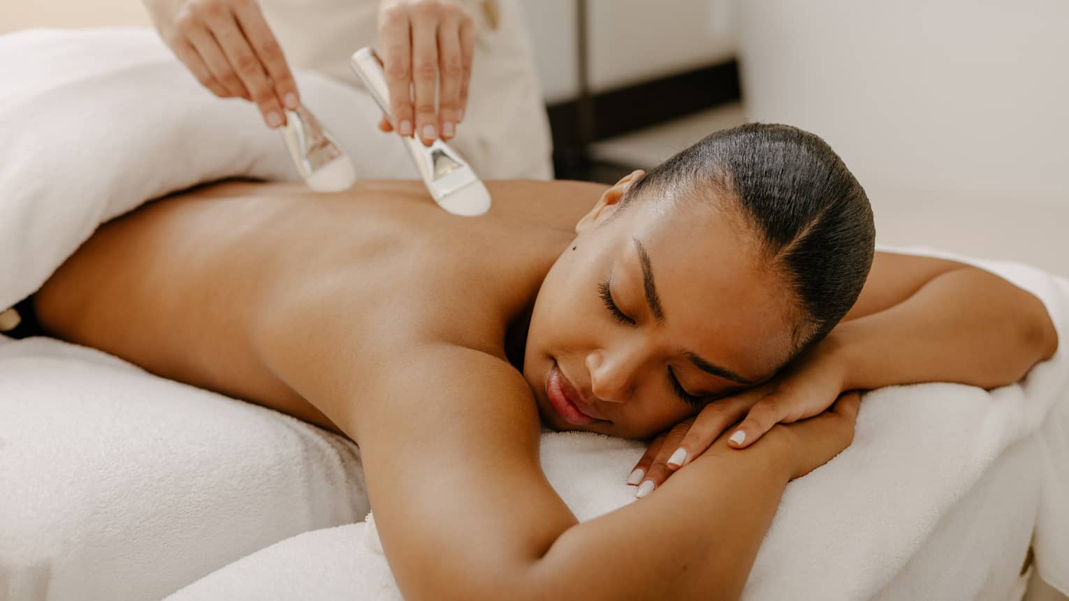 Two brushes massage a woman's back as she lay on a spa table covered with white linens