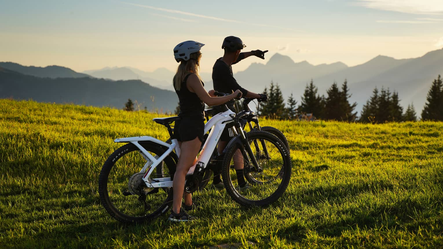 Two people riding fat bikes on meadow, one pointing to distant mountains
