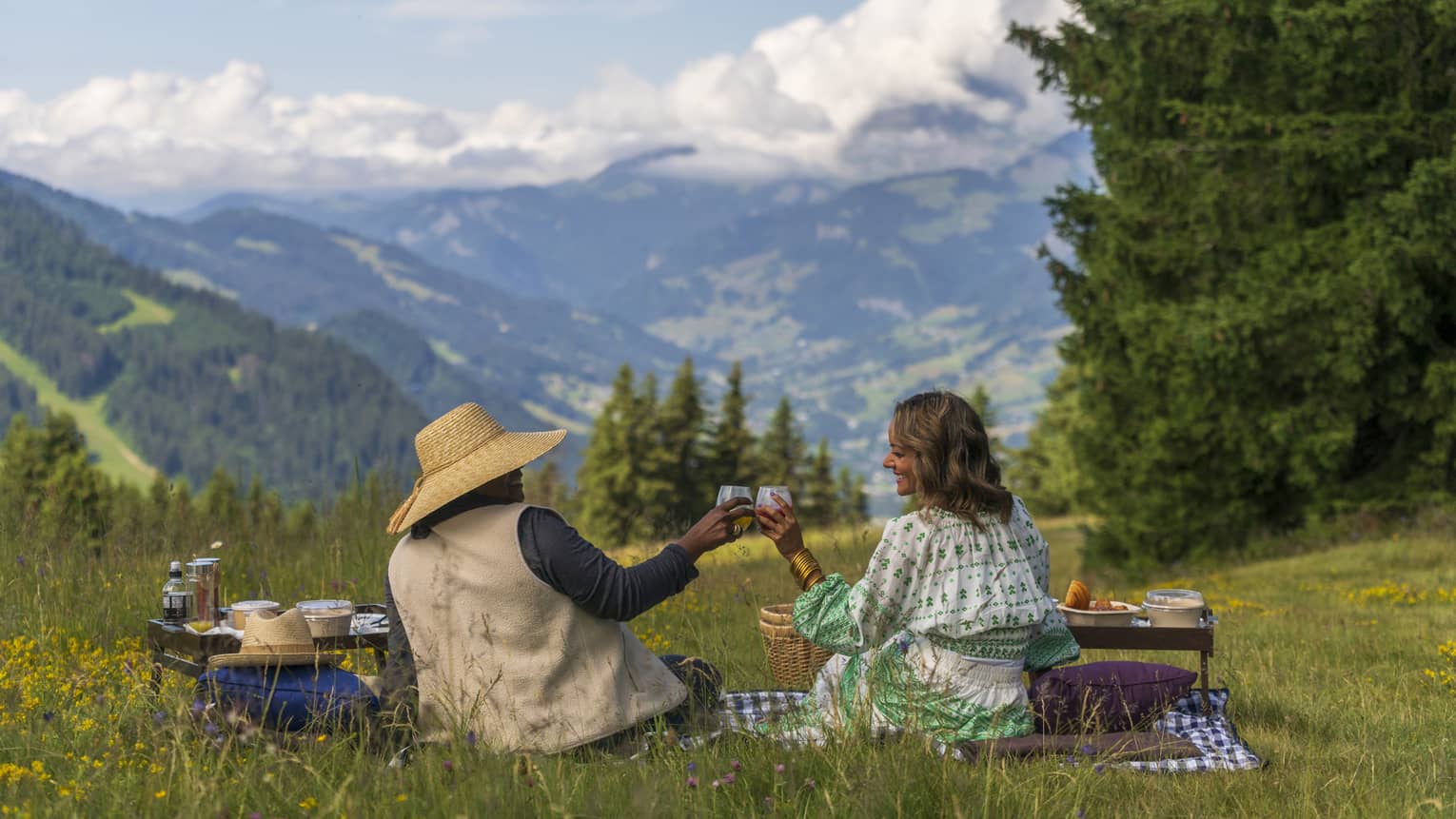 A couple sharing a picnic on a blanket in grass speckled with wildflowers, overlooking the rolling hills and forests.