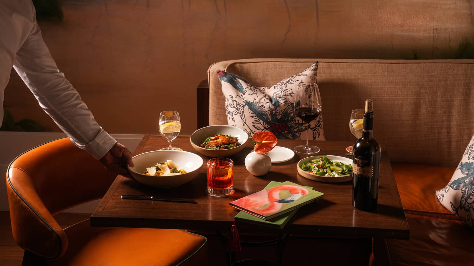 Server's hand places a bowl onto a rectangular wood dining table holding two other dishes, a bottle of wine, two water glasses, a wine glass and a cocktail in a rocks glass, with an orange leather chair and a beige-and-orange banquette nearby