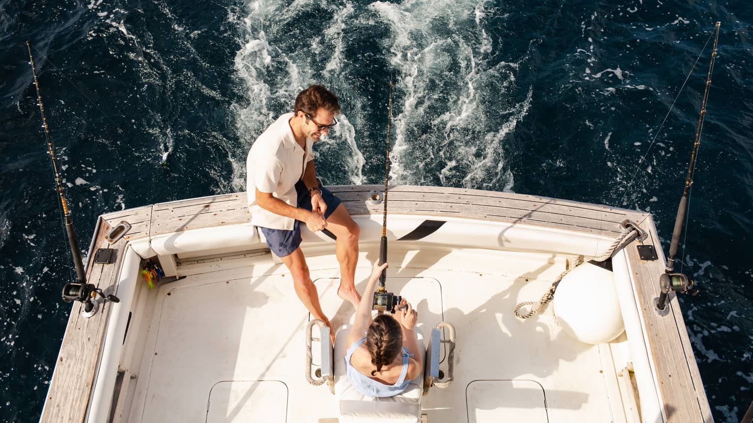 Two people sit at the back of a fishing boat, with one person holding a fishing rod and the other sitting close by on the edge of the boat